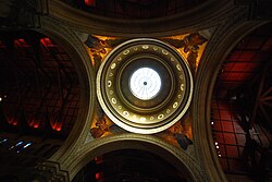 Looking upward, a view of the church's dome: at the top of the dome is a circular window or oculus. Beneath the skylight are several encircling bands decorated with mosaics including scrolling vine motif. The dome is supported on triangular pendentives, each decorated in mosaic with the figure of a standing angel with outstretched wings.