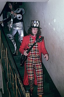 A man walks down a flight of stairs. He holds a guitar and wears a black hat studded with round metal mirrors. His waistcoat and trousers are tartan. His coat and shirt are plain red. His socks are striped.