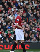 A side-on photograph of a man with red hair. He is wearing a red shirt, white shorts and black socks.