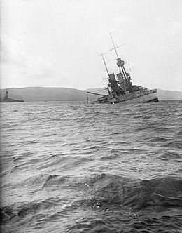 A large light gray battleship tilts back in choppy water as it slowly sinks.