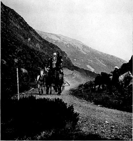 Several horses pulling a carriage up a hill on a dirt road surrounded by hills