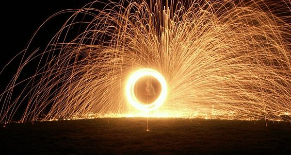 A fire performers spinning poi consisting of lit wire wool in chicken wire cages, dipped first in paraffin. Long-exposure photography captures the tra