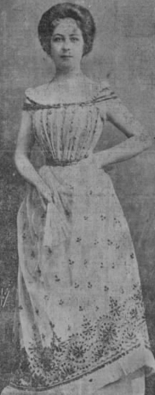 A young white woman wearing a gown with beetlewing trim. She is standing, one hand on hip, one hand holding fan. Her hair is in a bouffant updo.