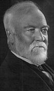 Bust portrait photograph of an old man, mostly balding with some whispy white hair on top, with a trimmed white goattee.