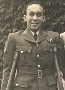 Black and white upper body photograph of a young adult black man wearing a British Royal Air Force uniform. He stands with the help of crutches visible under both his arms.