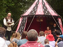 À la droite de la photo se trouve une tente dans laquelle sont assis un homme et une femme. À la gauche, un homme en habit d'époque fait la lecture debout devant des spectateurs assis.