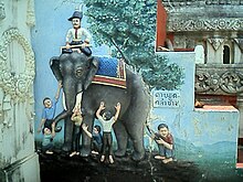 An einer Mauer (vielleicht in einem Tempel) ist das Gleichnis der Blinden mit dem Elefanten als Relief abgebildet. Die Darstellung könnte etwa einen Meter hoch sein; sie ist naturnah und sehr plastisch ausgeführt und in natürlichen Farben koloriert. Die Gruppe steht auf einer Anhöhe, hinter der sich eine weite, nur angedeutete Landschaft bis zum Horizont erstreckt. Der Himmel ist hellblau. Unmittelbar hinter dem Elefanten steht ein großer, sehr detailreich dargestellter Baum, rechts daneben befindet sich eine Tafel mit thailändischen Schriftzeichen. Der dunkelgraue Elefant steht mit dem Kopf nach links quer vor uns. Er hat den Kopf dem Betrachter zugewandt und scheint sich an dem Tun der Männer nicht zu stören. Über seinen Rücken ist eine Decke geworfen, sie zeigt ein blaues Rautenmuster und hat eine gelbbraune Bordüre. In seinem Nacken hockt im Schneidersitz ein Elefantenführer in schmucker Uniform mit dunkler Hose und weißem Hemd, er hat einen gezwirbelten Schnurrbart und trägt einen mit einer weißen Feder geschmückten blauen Hut. Er scheint nicht zu den Blinden zu gehören. Sechs blinde Männer stehen oder hocken um den Elefanten herum. Sie sind mit einfachen Hemden und Hosen bekleidet und barfuß. Sie betasten mit geschlossenen Augen ein Ohr, den Rüssel, einen Stoßzahn, einen Oberschenkel, den Bauch und den Schwanz des Tieres, welches dies ungerührt über sich ergehen lässt.