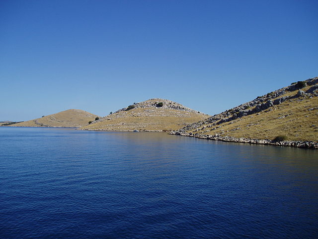 A photograph of a beige archipelago with a dark blue sky above fading to light blue on the horizon and a dark blue ocean in the foreground also fading to light blue at the coast