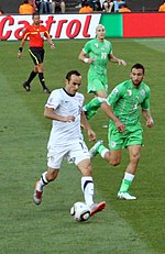 A man wearing a white shirt plays with a round ball at his feet, while being chased by two men in green shirts on a grass field.