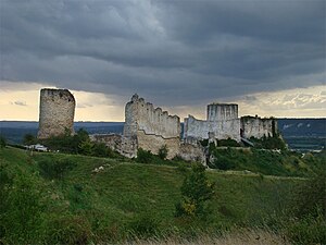 The ruins of a castle in grey limestone. It dominates the landscape. The castle's keep protrudes above the walls of the inner bailey; to the left a tower stands taller than the ruined walls.