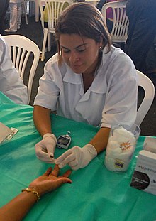 eine Krankenschwester führt eine Blutzuckermessung bei einem Patienten vor, beide sitzen an einem Tisch