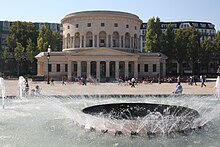 The rotunda can be seen in the center of the picture across a square with a fountain.  The building consists of a round structure surrounded by a colonnade with double columns, the entrance of which is a classic portico with eight columns and an empty tympanum.  Such porticos also lead off the rotunda to the left and right.