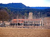 Foto eines Kirchturms und von Wirtschaftsgebäuden auf einem Feld;  im Hintergrund eine bewaldete und hügelige Landschaft.