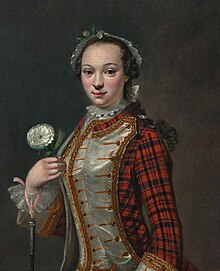 An young woman in a coat of predominantly-red tartan and white satin with gold braid, and a white headdress, with a white rose and what is probably a riding crop