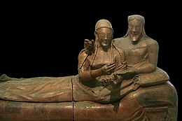 A sculpture of a woman and man reclining together on a couch, their upper bodies to the right and their legs to the left. There is a marked contrast between the high relief busts of their upper bodies and the very flattened lower bodies and legs. They have almond-shaped eyes and long braided hair, and are smiling widely. The man has a beard that is bobbed. The woman's hands are gesticulating in front of her as if she was holding something that is no longer there, or perhaps gesturing while speaking. The man has his right arm draped around the woman's shoulders in an intimate pose, and his right hand on her shoulder also appears to once have held some item. His left hand rests palm up in the crook of the woman's left elbow.