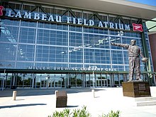 Photo of the side of Lambeau Field, with the words "Lambeau Field Atrium" on the side. Bob Harlan Plaza is located in front of the stadium. A small plaque highlighting Bob Harlan is located in the center of the plaza, with a statue of Curly Lambeau on the right.