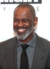 An image of McKnight wearing a gray suit and black tie while smiling to a camera in a red carpet event