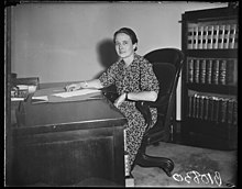 A white woman wearing a print dress, seated at a large desk