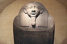 A dark stone Egyptian sarcophagus. The image shows the face of the sarcophagus in a relaxed position looking to the horizon. The sarcophagus shows left-to-right inscriptions in Phoenician on its lid.