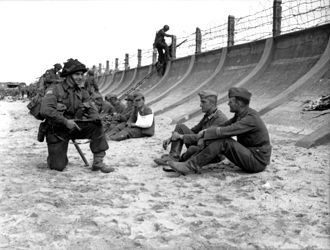 Juno Beach on D-Day, 1944. The barbed wire fence is crude and not very high. However, when combined with the steep, curving sea wall it slows down any attacker, giving time for the machinegun bunker (visible on the far left) to enfilade any attackers. Note the soldier in the background, forced to use a ladder. German POWs Juno Beach.jpg