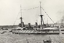 A World War I-era warship with tents erected over the fore and aft decks and flying flags from her rigging. Other warships, a small fortress and land are visible behind the ship and several small boats are visible in the foreground.