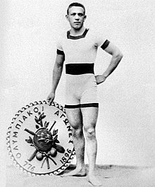 A man in swimming attire holding an emblem inscribed with "Olympic Games" in Greek lettering and the numbers 776 and 1896