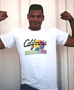 Ein hellhäutiger Schwarzer in einem weißen T-Shirt mit der Aufschrift "California" lächelt und streckt seine Arme für die Kamera.