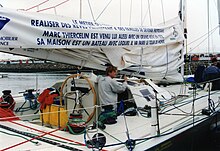 Vue de trois quarts arrière, le cockpit d'un voilier, avec une barre à roue et un homme vu de profil.