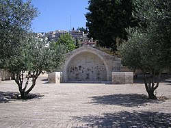 A structure of white stone containing an arch is seen in a plaza of similar stone. Two short trees are shown in the foreground.