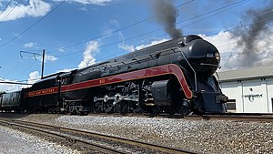 A posed side view of a large 4-8-4 streamlined steam locomotive with two tenders. The overhead power lines and a building are in the background.