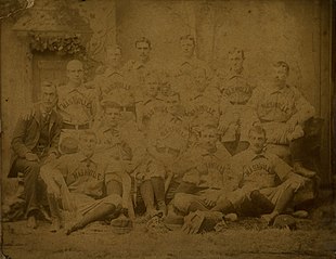 A sepia photograph of 14 men arranged in two rows, standing and sitting. One is wearing a suit while the rest are wearing light baseball uniforms with 