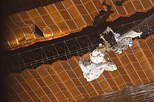To sorte og orange solpaneler, vist ujævne og med en stor flænge synlig.  Et besætningsmedlem i en rumdragt, fastgjort til enden af ​​en robotarm, holder et gitterværk mellem to solsejl.