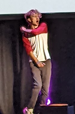 Young man with curly hair dressed casually in multi coloured tee shirt gyrating on a theatre stage addressing the audience by way of a headset microphone.
