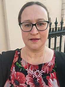 Laurence Totelin, a white woman, wears a flowery dress and a black cardigan, as well as a necklace showing showing a molecule and black glasses. she is lightly made up.