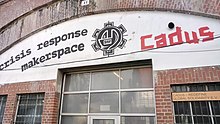 Eingangsbereich eines Backsteingebäudes mit großem Schriftzug „Cadus Crisis Response Maker Space“ und Logo über den Flügeltüren