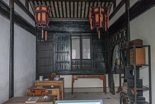 Fotografie nevelké místnosti, s oknem uprostřed zadní dřevěné stěny a úzkým stolkem a židlí pod oknem, dále vybavené třemi stolky po levé straně, skříní při přední části pravé stěny, za níž je průchod do sousední místnosti, z plochého dřevěného stropu visí dva zdobné lampióny
