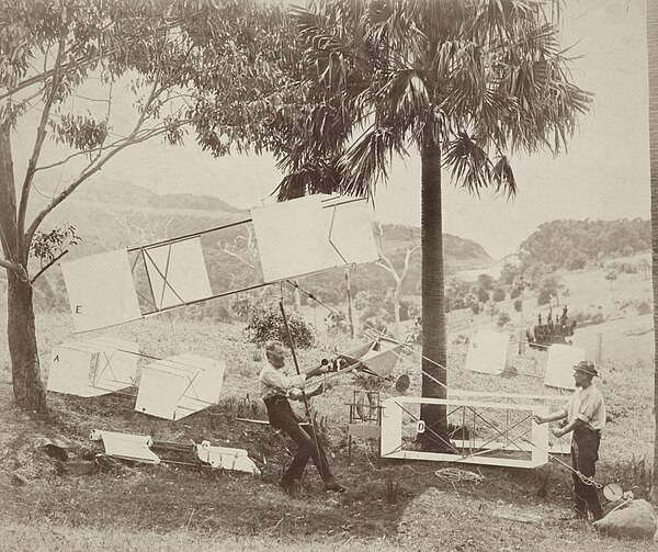 Hargrave (seated) and Swain demonstrate the manlift kites (labelled A, B, D, & E), sling seat and spring balance in the parkland behind Stanwell Park 