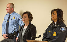 A police officer in uniform, a mayor in a black blazer, and a police chief in a dark blue police jacket stand before a podium.