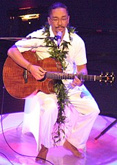 A man in white clothing on a stage, seated and holding a guitar