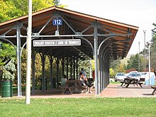 Long and narrow wooden-roofed canopy, supported by two rows of ten metal pillars, with wooden sign reading 'trolley station' and picnic tables underneath