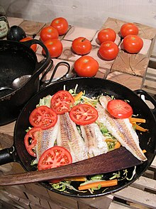 Fillets of fish on a bed of shredded vegetables cooking in a frying pan, partially covered by sliced tomatoes.