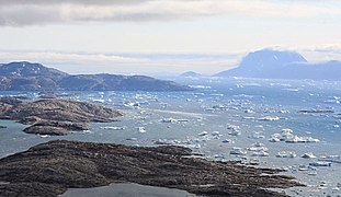 Photographie en couleurs d'un fjord aux eaux parsemées de petits blocs d'icebergs et bordé de côtes échancrées à gauche et d'île à droite, en arrière-plan.
