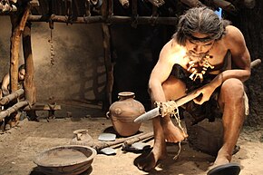 A shirtless man with long hair wearing a bone necklace, twirling twine around a wooden stick while surrounded by several clay pots