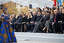 Angélique Kidjo de face, en pagne wax de couleur dominante bleue, main gauche sur le ventre, avec un micro dans sa main droite devant des chefs d'État assis sous un hangar.