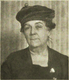 B&W portrait photo of a woman with her hair in an up-do, wearing a dark jacket with square neckline, and a dark cloche hat.