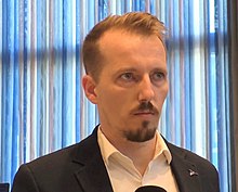 Caucasian man with dark blond hair and facial hair in a suit standing indoors