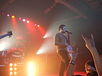 A Caucasian man sings into a microphone on a brightly lit stage. In the background, another man plays the drums.