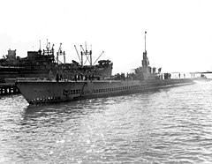 A long low-slung naval ship is entering a dock. Its deck is lined with sailors.