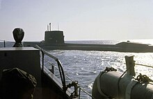 Farbfoto, das die Backbordseite eines großflächigen Atom-U-Bootes zeigt, das vom Bug eines eskortierenden Überwasserschiffs aufgenommen wurde, als das U-Boot den Hafen verlässt.