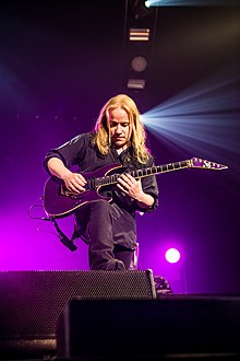 Kytarista skupiny Emppu Vuorinen během koncertu hrající kytarové sólo ve vyšších tónech. Fialovou kytaru značky ESP EV-1 má opřenou o pravou pokrčenou nohou.